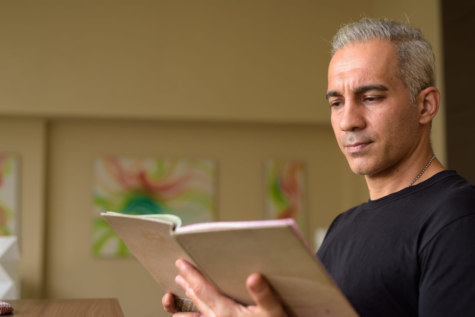 Homem sentado lendo um livro em ambiente doméstico, com quadros coloridos ao fundo.