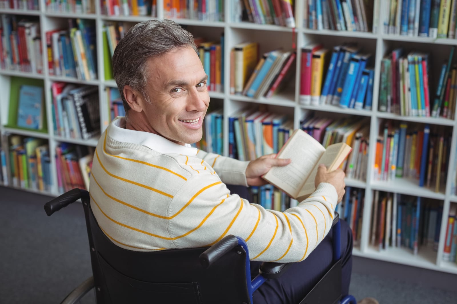 Homem em cadeira de rodas sorri segurando um livro aberto diante de estantes coloridas em uma biblioteca.