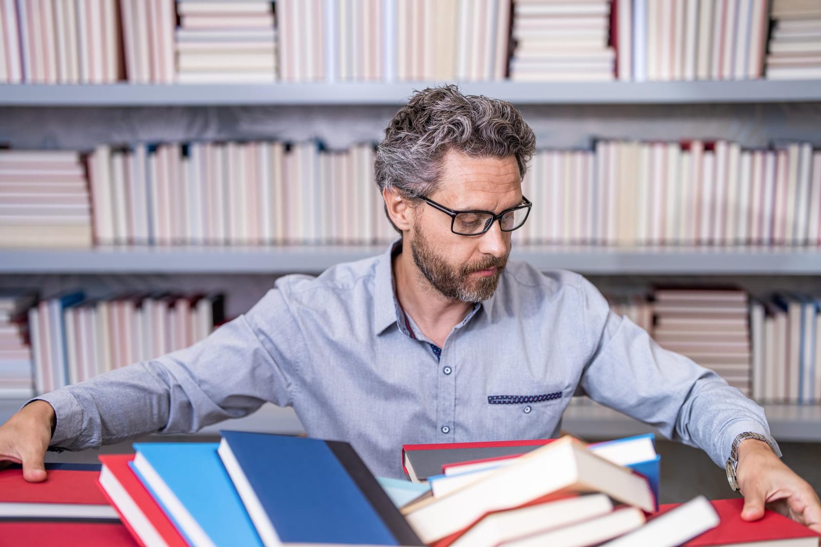 Homem organiza uma pilha de livros de capas coloridas sobre uma mesa, diante de estantes de biblioteca.